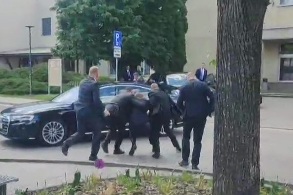 A white man is carried to a black car by two other people. Two people stand on either side of them.