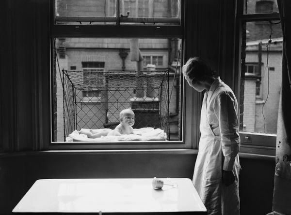 A black and white photo shows a nurse wearing a white coat looking at a baby in a wire cage attached to a window with blankets covering the bottom looking at them. 