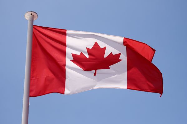 A red and white flag with a red leaf in the middle is flying in a blue sky.