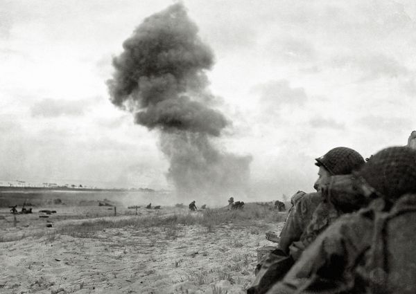 Two soldiers stand near the right corner of an image that showed a beach. Smoke comes up from the middle of the beach, where you can see other soldiers.