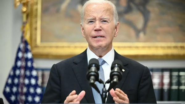 A white man wearing a black suit stands behind a podium that has a microphone on it.