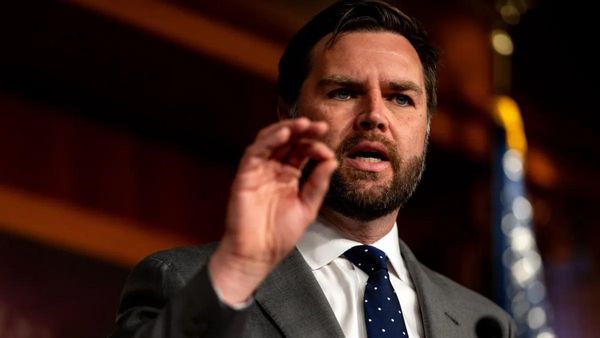 A white man wearing a gray suit speaks with the pointer finger and thumb of his left hand touching.
