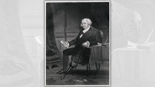 A white man sits in a chair holding a document in his left hand.