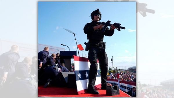 A person holding a rifle and wearing a black uniform is standing on a platform. You can see a crowd of people distantly to the side, as well as other people huddled near a podium.