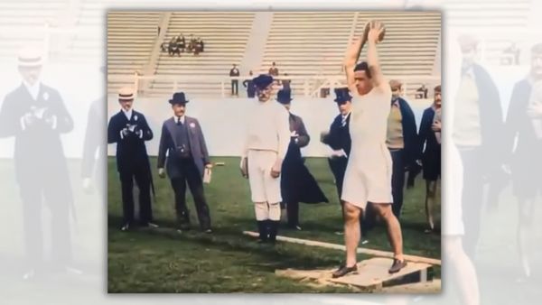 A white man holds a ball over his head standing on a raised piece of wood. Several people wearing suits surround him, and a small crowd sits in the bleachers.