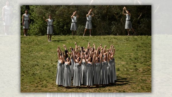 A group of women are centered in the grass. Above them, five men are standing, some with their arms stretched above their heads.