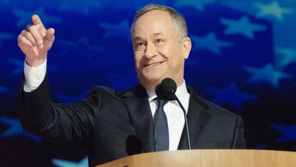A white man wearing a black suit holds his left hand out while standing behind a podium with a microphone attached.