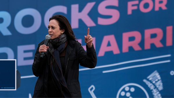 A Black Asian woman wearing all black clothing holds a microphone in her left hand while holding up her right pointer finger. 