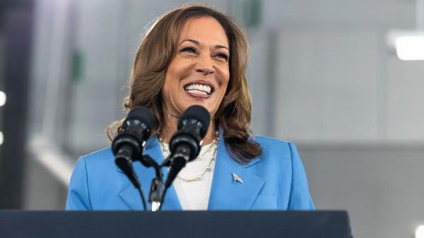 A Black Asian woman wearing a blue suit stands smiling behind a podium that has a microphone attached.