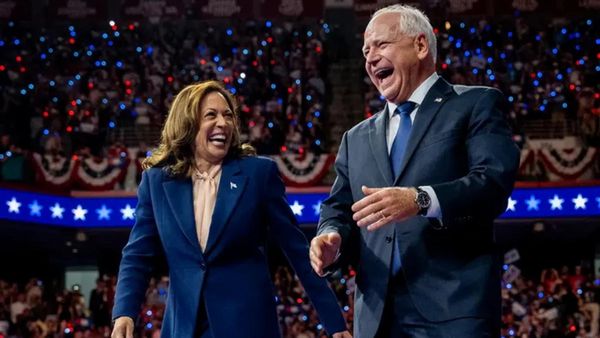 A Black Asian woman in a blue suit stands next to a white man wearing a gray blue suit. Both look happy and are standing in front of a large crowd.