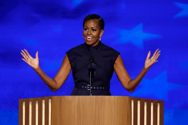 A Black woman stands at a podium that has a microphone on it with her arms outstretched.