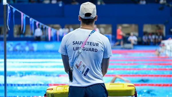 A person wearing a shirt that says "SAUVETEUR LIFEGUARD" on it stands near a pool with several lanes.