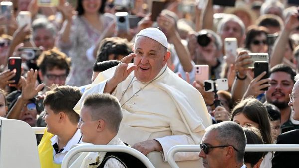 A white man holds his left arm near his face. A huge crowd surrounds him, many people holding phones up like they are recording him.
