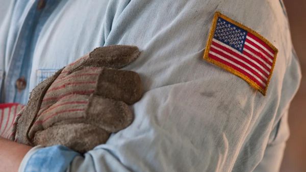 A person wearing a blue button down shirt with an American flag patch on it has their arms crossed. One hand is covered in a brown glove.