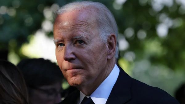 A white man wearing a black suit looks towards the camera. His face is partly shrouded by shadows.