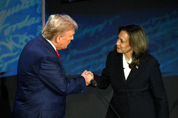 A white man wearing a blue suit shakes hands with a Black Asian woman wearing a black suit.