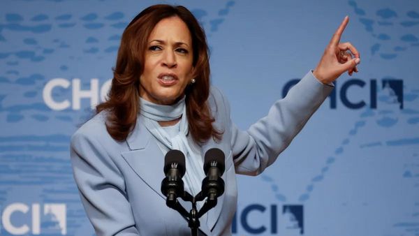 A Black South Asian woman wearing a light blue suit and scarf holds the pointer finger of her right hand up while standing at a podium.