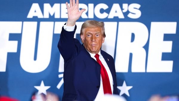 A white man wearing a blue suit raises his left hand in greeting. Large white letters that say "AMERICA'S FUTURE" are behind him.