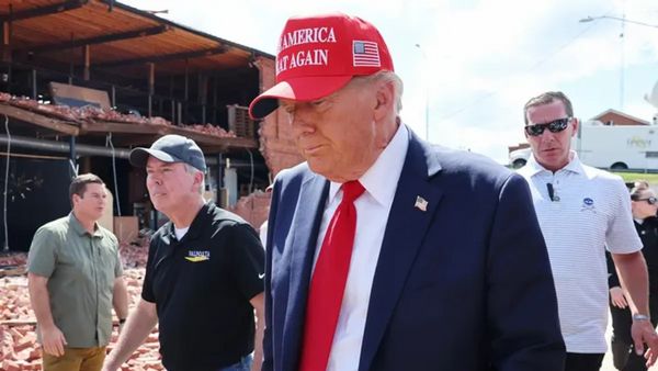 A white man wearing a red MAKE AMERICA GREAT AGAIN hat and blue suit looks down. The American flag can be seen on the side of the hat. Several people can be seen on either side of him.