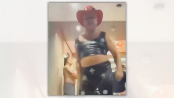 A white man wearing a black crop top and pants with a red cowboy hat is blurrily pictured.
