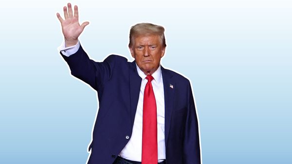 A white man wearing a blue suit raises his hand in what looks like a greeting over a light blue background.
