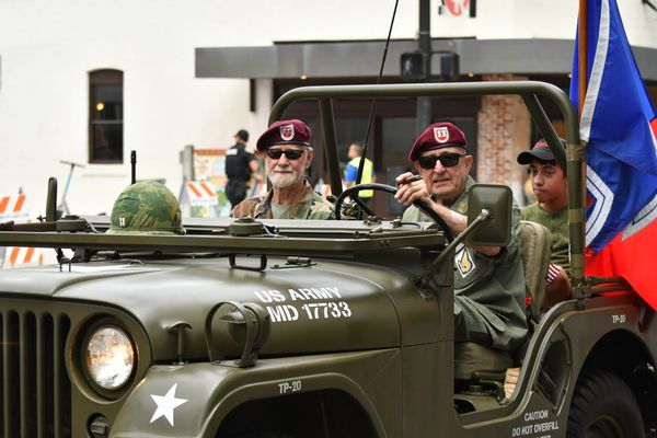 Two people sit in the front of a vehicle that says "US ARMY MD 17733." A person sits behind them, with a flag faintly seen towards the back as well.