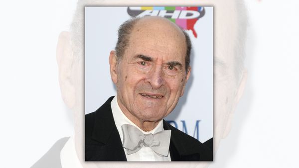 A white man wearing a black and white suit looks toward the camera.