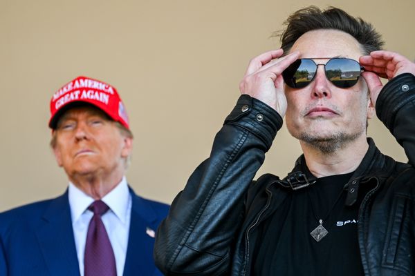 U.S. President Donald Trump wearing a red "MAKE AMERICA GREAT AGAIN" hat and a blue suit is in the background on the left of the photo. Elon Musk, a white man with short brown hair wearing a black leather jacket and aviator sunglasses, puts his sunglasses on in the foreground of the photo on the right. 