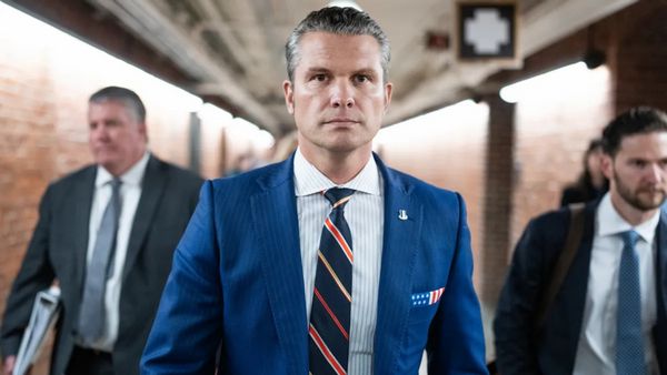 A white man wearing a blue suit walks down what appears to be a hall. Several people can be seen behind him.