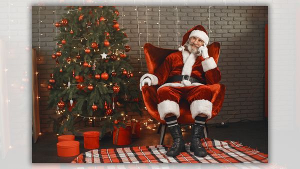 A white man sitting in a red chair wearing a red hat and sui is pictured next to a tree decorated with ornaments with presents underneath it.