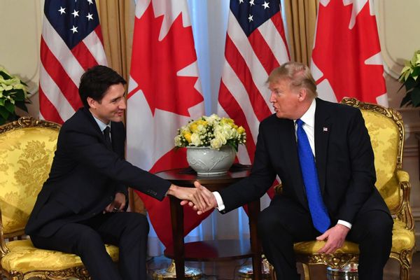 Prime Minister Justin Trudeau of Canada (left) shakes hands with then-U.S. President and now U.S. President-elect Donald Trump. Both men are wearing suits and sitting in ornate yellow chairs. Canadian and American flags fill the backdrop. 