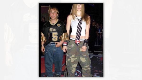 A white man and woman stand next to each other. The man on the left is wearing a jersey with "5" on it, while the woman is wearing a white tank top and cargo pants. A tie is draped around her neck.