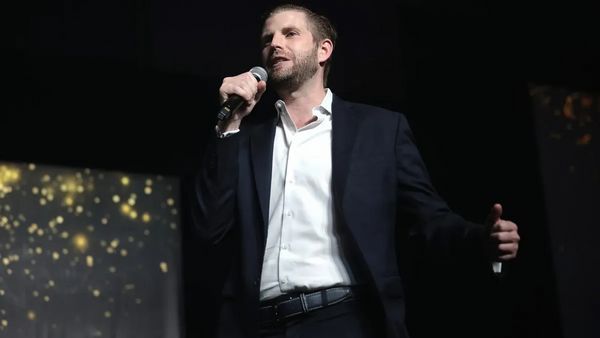 A white man wearing a dark blue suit jacket and white shirt speaks into a microphone.