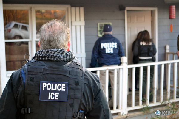 The back of an ICE official wearing a bulletproof vest with ICE written on it. In the background, another ICE official is knocking on a door. 