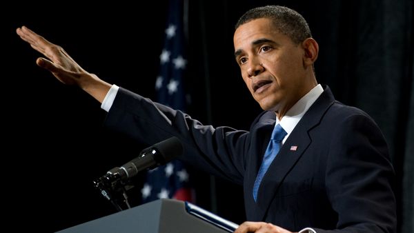 A Black man wearing a blue suit stands at a podium. His left arm is up in a sharp angle.