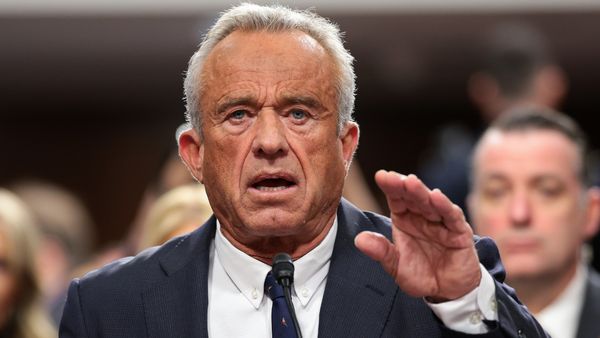 A white man wearing a gray suit holds up his right hand while speaking.