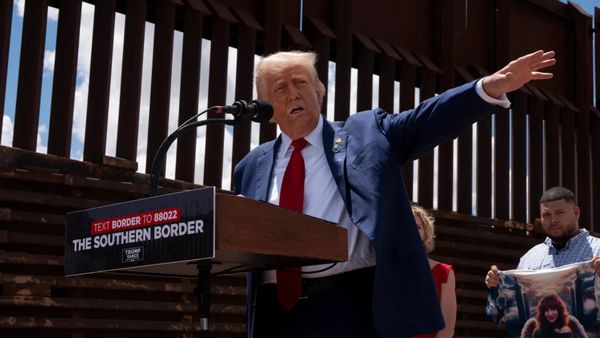 A white man wearing a blue suit outstretches his arm while standing behind a podium. A wall can be seen behind him.