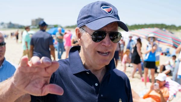 A white man wears sunglasses and a blue hat at a beach. Behind him, you can see a crowd of people.