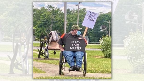 A white man sitting in a wheelchair holds a sign in his lap that says BLACK LIVES MATTER. A sign attached to his wheelchair says, "I'M SORRY I'M LATE. I HAD A LOT TO LEARN."
