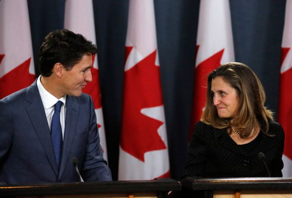 A tall white man with brown hair in a blue suit and tie talks to a white woman with blonde hair in a black jacket with small white dots. They stand in front of a wall of Canadian flags -- white and red vertical stripes with a red maple leaf emblazoned in the middle.