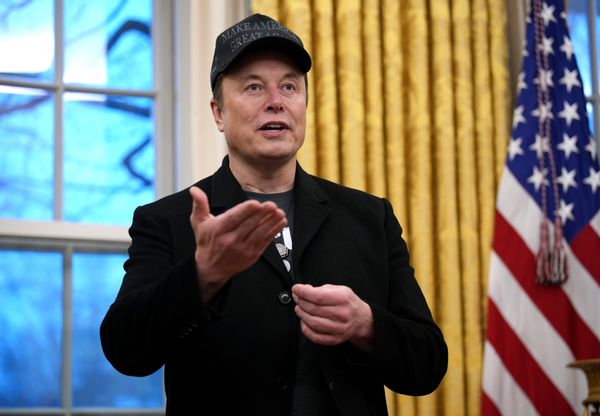 A white man wearing a black jacket and MAKE AMERICA GREAT AGAIN hat stands with his left hand flat.