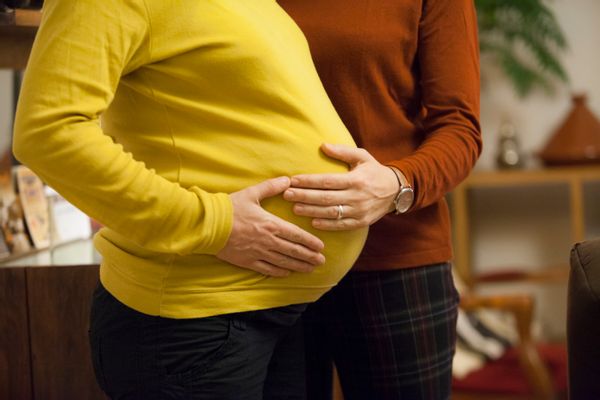 A pregnant person in a yellow long-sleeved shirt stands in profile (their head is not in view). Another person in a long-sleeved rust orange shirt wearing a watch and with a ring on their ring finger places their hand on the pregnant person's belly.