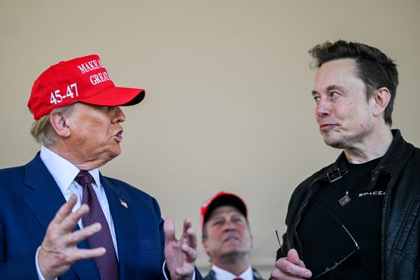 U.S. President Donald Trump, a white man with a dark tan in a blue suit wearing a red "Make America Great Again" baseball cap, talks to Elon Musk, a white man with short brown hair and a five o'clock shadow wearing all-black.