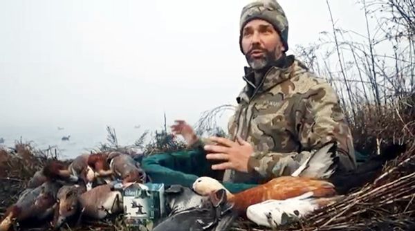A white man is pictured looking towards a camera wearing a camoflauge hat and jacket. Ducks surround him. He appears to be sitting outside near a body of water.