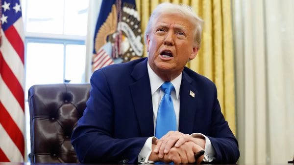 A white man wearing a blue suit sits at a desk with his right hand held over the left.