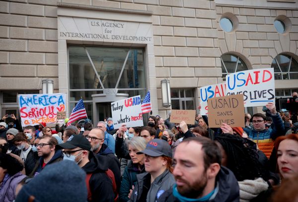 A crowd of people, many of whom are holding signs that say things like "USAID saves lives" and "America, seriously? A dictator?" outside of a building which is labeled "U.S. Agency for International Development"