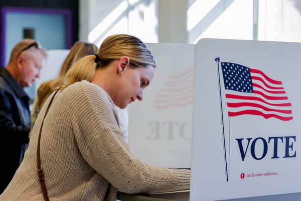 A white woman with blonde hair in a ponytail wearing a beige sweater leans over a document behind a large white privacy folder that says "VOTE" with the U.S. flag on it. 