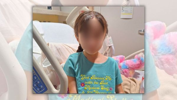 A picture of a young girl, with her face blurred out, in front of a hospital bed. She has a single brown braid and is wearing a light blue t-shirt that says "God blessed me with the best medical staff and second chance at.." (the t-shirt is cut off in the image there) 
