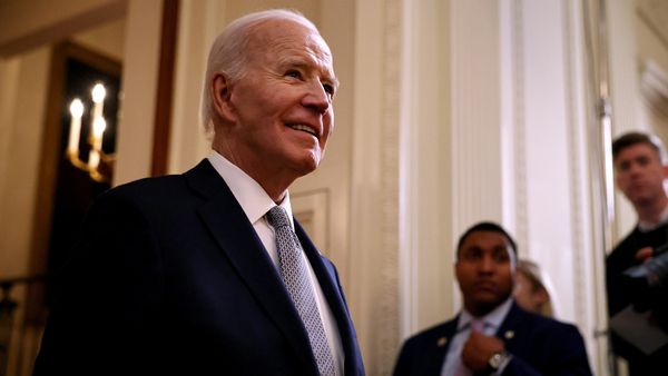A white man wearing a black suit stands in a room smiling. Two other people can be seen by the side.