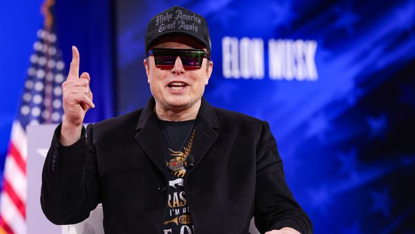 A white man wearing a black jacket and sunglasses speaks with the index finger of his left hand raised,